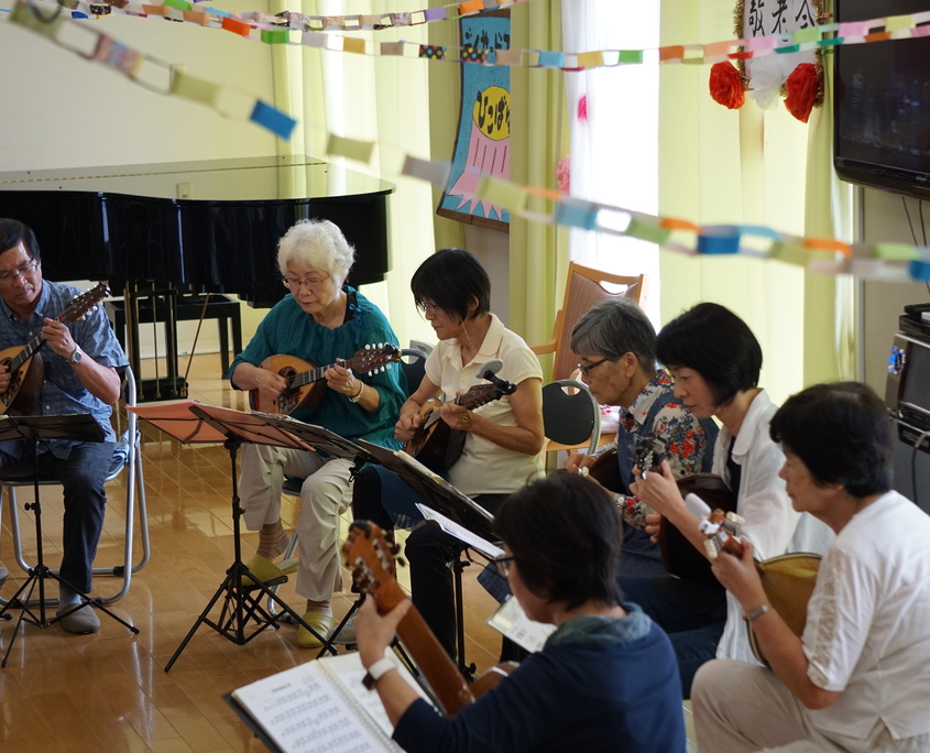 介護施設でのギター演奏レクリエーション。複数の入居者と職員が楽器を持ち音楽活動に参加している。